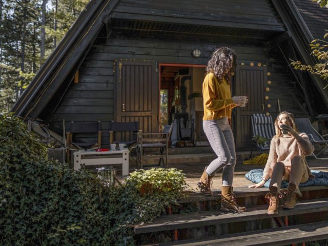 Two beautiful young female friends sitting on stairs on porch in front of log cabin, talking and drinking coffee