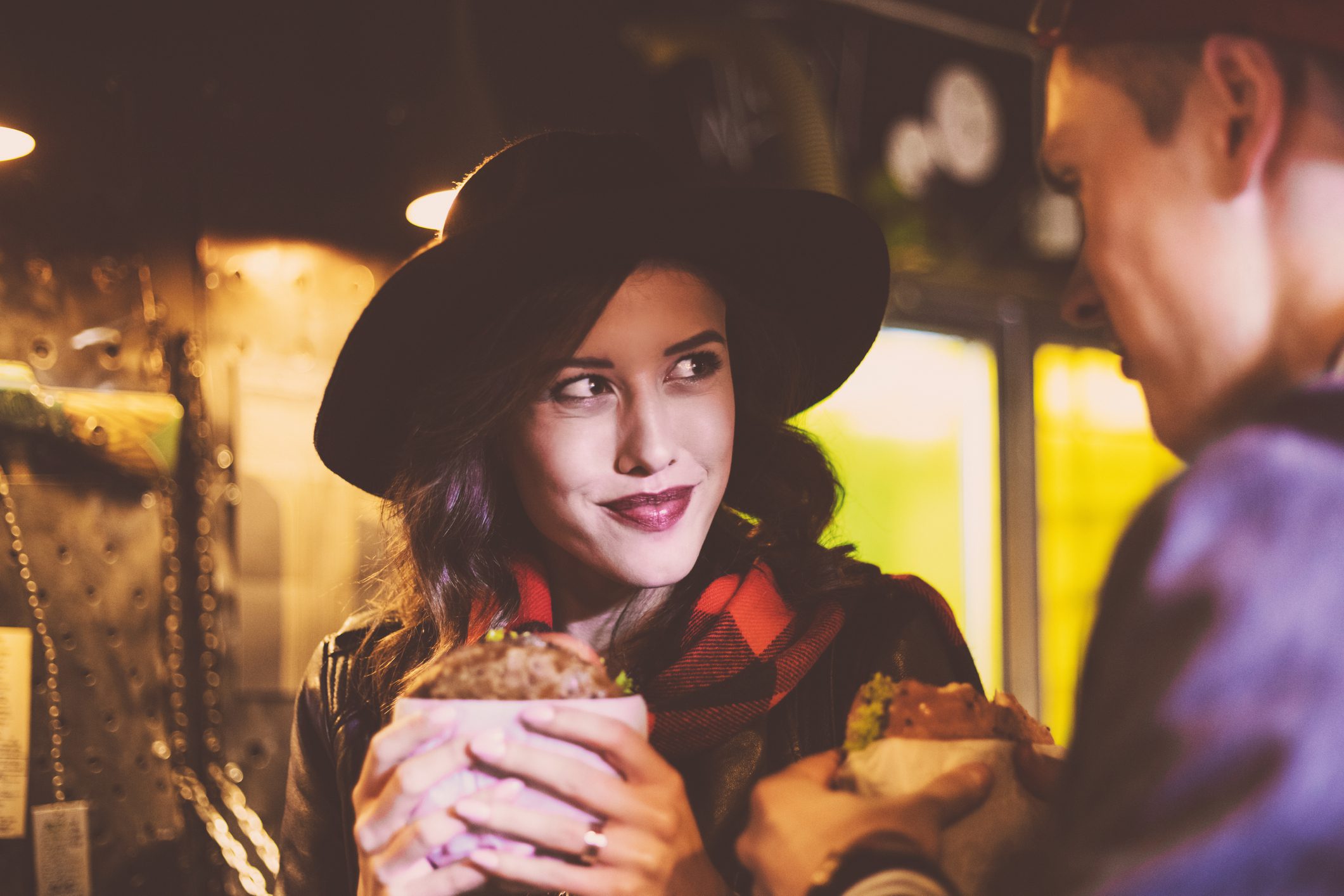 Beautiful young woman wearing black hat eating burger in the pub. Close up of face. Dark tone.